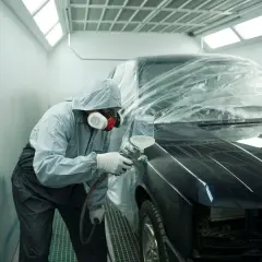 Tecnico in tuta protettiva vernicia un'auto in cabina. Il costo del ripristino trasparente dell'auto è visibile nel processo.