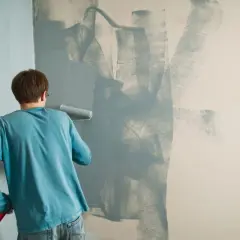 Un uomo con una maglietta blu sta dipingendo un muro con un rullo. La pittura si stacca dal muro in alcune zone, creando un effetto astratto.