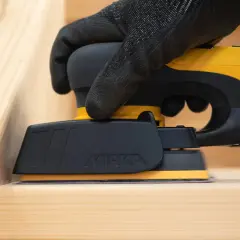 Mano guantata utilizza levigatrice elettrica per carteggiare legno grezzo, preparando la superficie per la finitura.