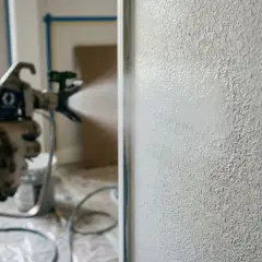 Mano guantata usa pistola a spruzzo per dare finitura a muro. Meglio rullo o pistola a spruzzo? La pistola offre un'applicazione uniforme.