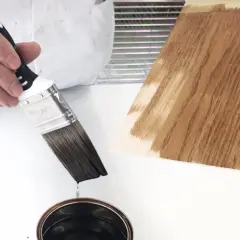 Pennello intinto in una latta di vernice scura, pronto per essere applicato su un campione di legno. Dopo l'impregnante, si mette la finitura.