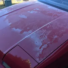 Il cofano di un'auto rossa presenta il trasparente rovinato, con chiazze bianche che indicano la necessità di capire come togliere il trasparente dalla carrozzeria.