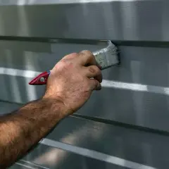 Mano che applica vernice grigia su una superficie a coste, probabilmente per verniciare alluminio e proteggerlo.