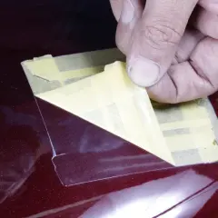 Una mano rimuove nastro adesivo da una superficie lucida color bordeaux. Il trasparente sulla vernice serve a proteggerla da graffi e usura, mantenendone la brillantezza.