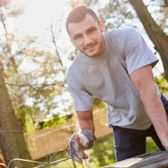 Uomo sorridente con guanti da giardinaggio lavora a un tavolo da giardino fai da te, immerso nel verde.