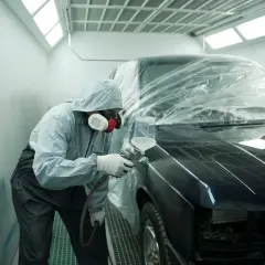 Un operaio in tuta protettiva e maschera spruzza vernice su un'auto scura, parzialmente coperta da un telo trasparente. L'auto sembra rovinata, in fase di restauro.