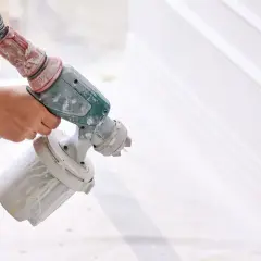 Mano che tiene un aerografo per verniciare con compressore, creando un effetto liscio su una superficie bianca.
