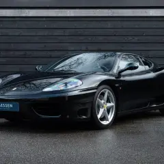 Una splendida auto nera, una Ferrari 360 Modena, parcheggiata su asfalto bagnato davanti a un muro di legno scuro.