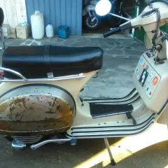 Vespa color crema con evidente effetto ruggine sul parafango posteriore. La sella è nera, con portapacchi cromato.