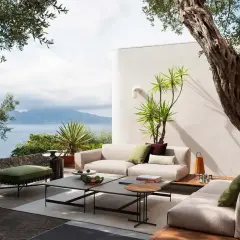 Arredare terrazzo con divani chiari, tavolini grigi e un pouf verde, con vista mare e scultura colorata.