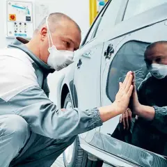 Un operaio con mascherina controlla la carrozzeria lucida di un'auto, pronto per carteggiare auto e rifinire la verniciatura.