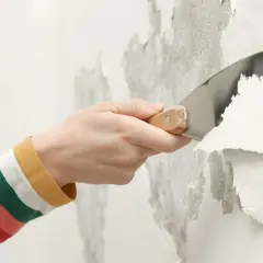 Mano con spatola rimuove vernice vecchia scrostata, preparando il muro per un nuovo strato, forse antiruggine.