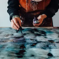 Mani che creano un'opera d'arte con colori acrilici su plastica, gocciolando inchiostro viola su una superficie texturizzata blu e oro.