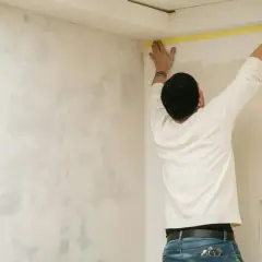 Uomo con cappello nero applica nastro giallo al soffitto, preparando la parete con un effetto jeans.