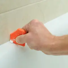 Mano usa spatola arancione per rifinire silicone, un lavoro antisilicone fai da te per sigillare la vasca.
