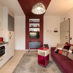 Soggiorno con divano bordeaux, tappeto grigio e una parete bordeaux e tortora che incornicia una nicchia con TV e mensole.