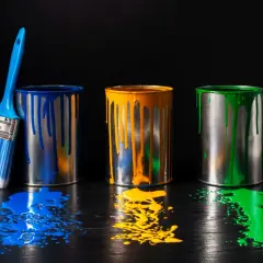 Tre barattoli di vernice (blu, giallo, verde) con gocce e schizzi su un tavolo scuro. Un pennello blu accanto. Il solvente cos'è?