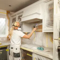 Donna con maschera protettiva effettua un cambio colore ai mobili della cucina con uno spruzzatore di vernice.