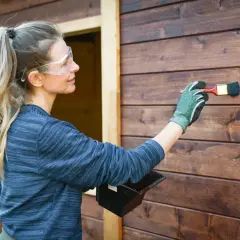 Donna con occhiali protettivi applica una vernice scura su una parete di legno, un tipo di pittura che protegge e abbellisce.