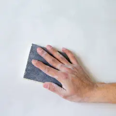 Mano che carteggia una parete bianca, un gesto che illustra il significato di carteggiare per levigare e preparare le superfici.