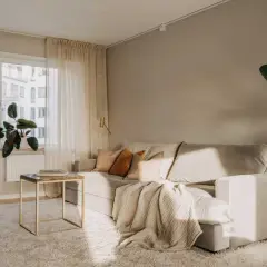 Un accogliente soggiorno con divano chiaro, cuscini color terra e una coperta morbida. Il tavolino dorato e la pianta verde aggiungono un tocco di stile. Pareti color marrone chiaro.