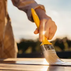 Mano che stende vernici poliuretaniche bicomponenti su un tavolo di legno al tramonto, per una finitura protettiva e duratura.