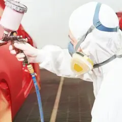 Persona in tuta protettiva e maschera effettua verniciare auto fai da te con pistola a spruzzo su un'auto rossa.
