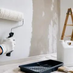 Mano guantata stende la primer vernice con un rullo su un muro grezzo. Scala, secchio e pennello a terra.