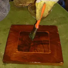 Mano guantata applica liquido con pennello per sverniciare mobile legno. Accanto, paglietta metallica e spazzola.