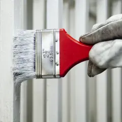 Mano guantata applica vernici industriali bianche su una ringhiera metallica con un pennello.