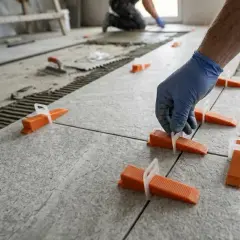 Mano con guanto blu che inserisce cunei arancioni per la posa piastrelle fai da te. Pavimento in fase di realizzazione.