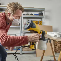 Uomo con mascherina usa un attrezzo per pitturare a spruzzo un telaio bianco su un tavolo da lavoro.