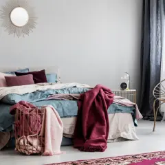 Letto con coperte color borgogna e cuscini, un cesto dorato con plaid rosa e una sedia dorata con cuscino turchese.
