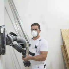 Operaio con maschera e occhiali sta per carteggiare muro senza fare polvere, usando un aspiratore integrato.