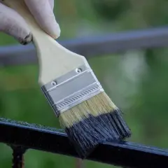 Mano con guanto bianco che usa un pennello per applicare vernice scura su una ringhiera, un passo per come sverniciare una ringhiera.