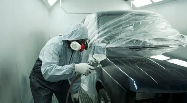 Tecnico in tuta protettiva vernicia un'auto in cabina. Il costo del ripristino trasparente dell'auto è visibile nel processo.