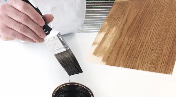 Pennello intinto in una latta di vernice scura, pronto per essere applicato su un campione di legno. Dopo l'impregnante, si mette la finitura.
