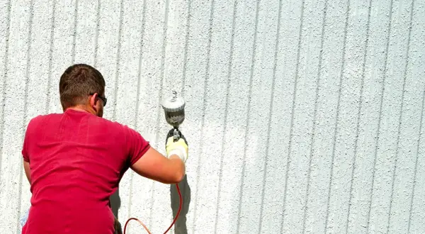 Uomo con maglietta rossa usa uno spruzzatore per verniciare una parete esterna. Ottimi trucchi per verniciare a spruzzo per un lavoro uniforme.