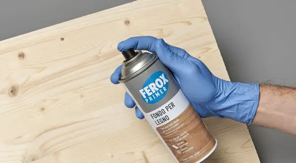 Mano con guanto blu tiene bomboletta spray "Ferox Primer Fondo per Legno", pronta per preparare la superficie.