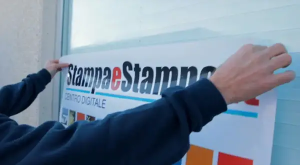 Mani applicano un grande adesivo con logo "Stampa e Stampe Centro Digitale" su una superficie, mostrando come applicare un adesivo senza bolle per un risultato perfetto.