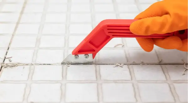Mano con guanto arancione usa uno strumento rosso per rimuovere lo stucco tra le piastrelle, un passo fondamentale per capire come togliere le piastrelle dal muro.