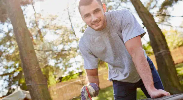 Uomo sorridente con guanti da giardinaggio lavora a un tavolo da giardino fai da te, immerso nel verde.