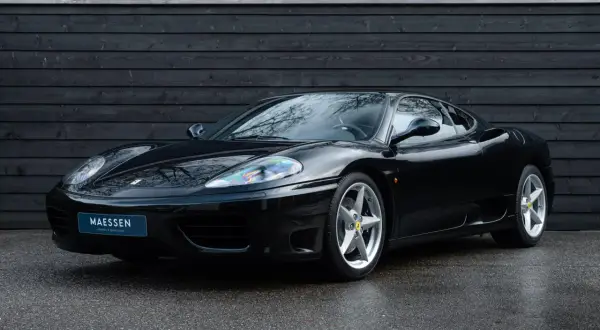 Una splendida auto nera, una Ferrari 360 Modena, parcheggiata su asfalto bagnato davanti a un muro di legno scuro.