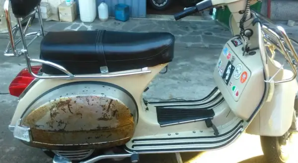 Vespa color crema con evidente effetto ruggine sul parafango posteriore. La sella è nera, con portapacchi cromato.