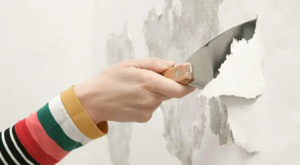 Mano con spatola rimuove vernice vecchia scrostata, preparando il muro per un nuovo strato, forse antiruggine.