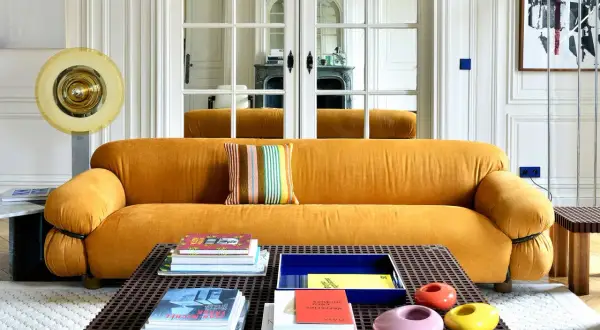 Divano senape con cuscino a righe colorate. Tavolino con libri e oggetti decorativi. Abbinamenti moderni e audaci.