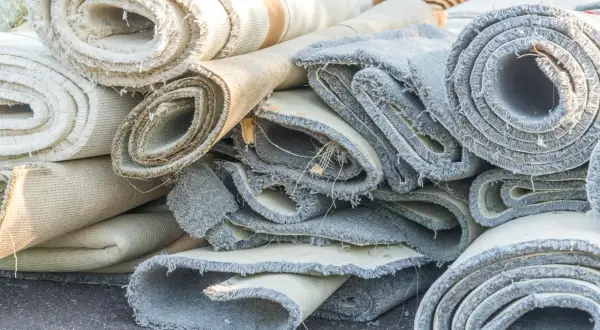 Rotoli di moquette accatastati, pronti per essere rimossi. Si vedono i bordi sfilacciati e i diversi strati dei tappeti.