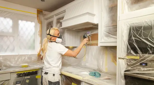Donna con maschera protettiva effettua un cambio colore ai mobili della cucina con uno spruzzatore di vernice.