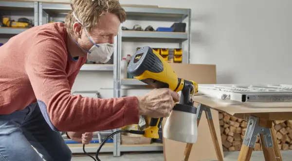Uomo con mascherina usa un attrezzo per pitturare a spruzzo un telaio bianco su un tavolo da lavoro.