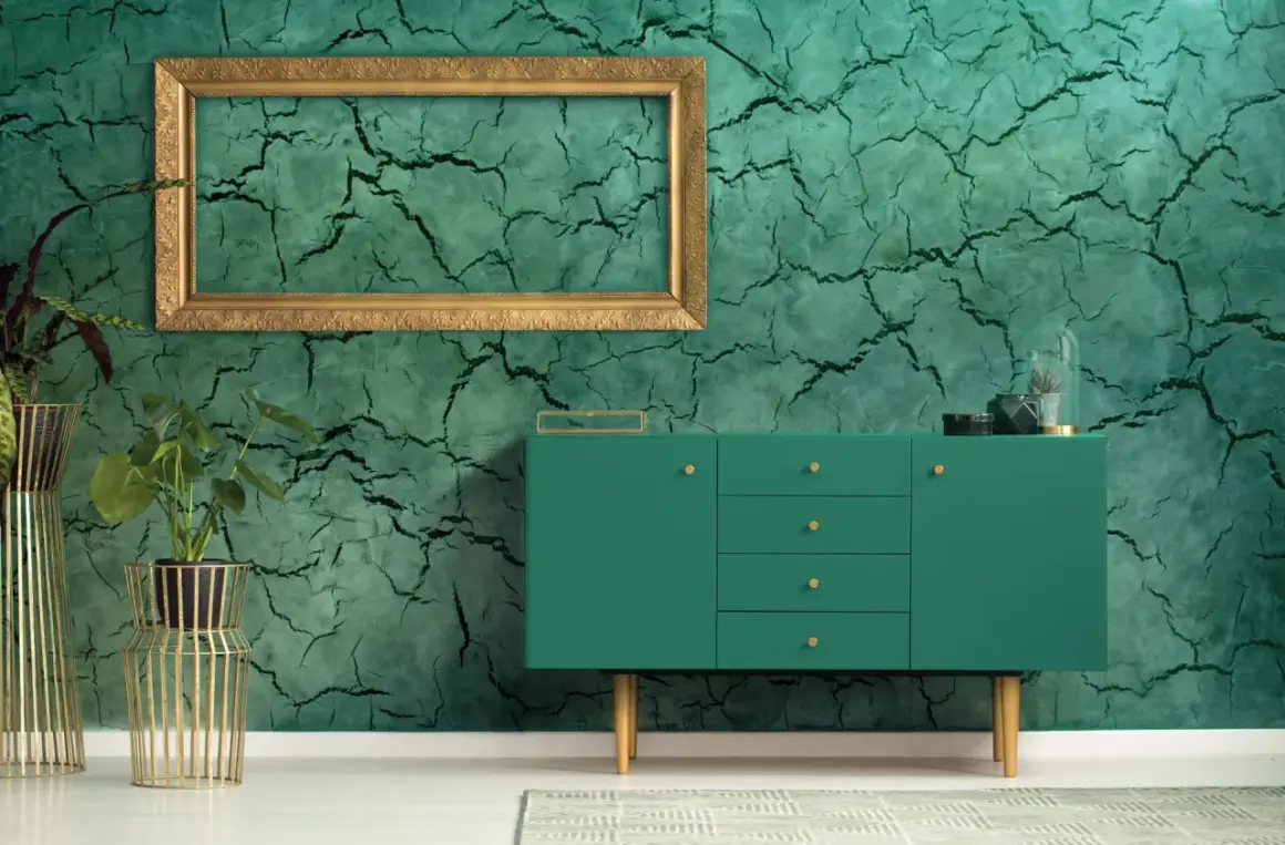 Parete con finitura in stucco materico verde intenso, incorniciata da un elegante mobile verde e piante in vasi dorati.